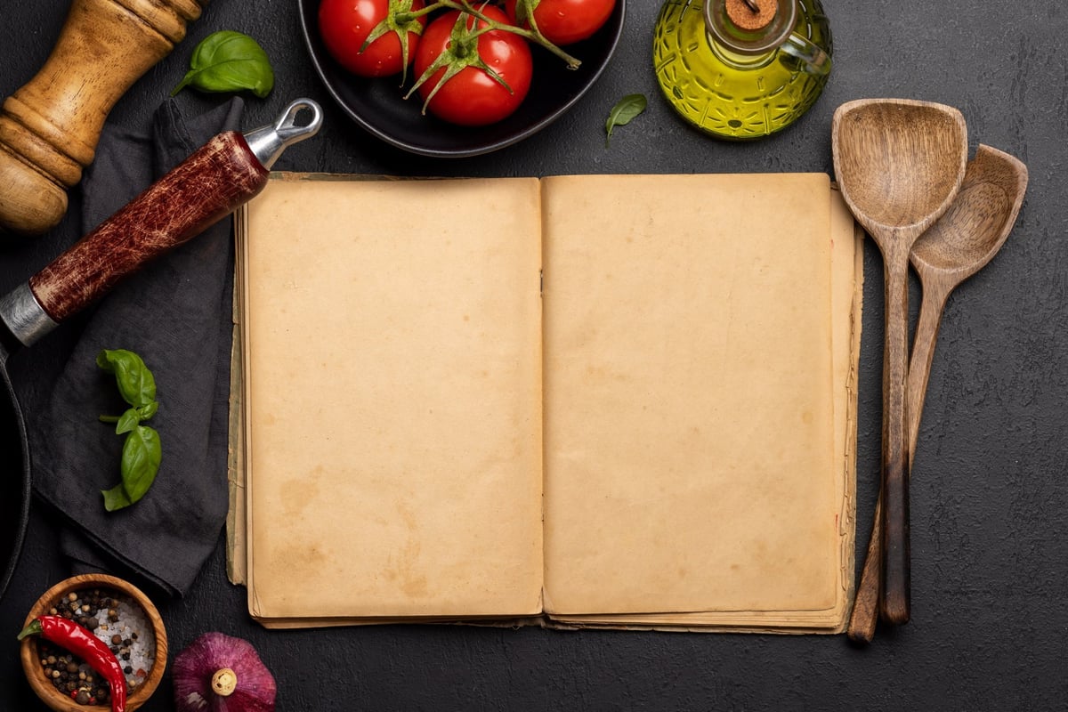 Top-down view of a kitchen table with ingredients, utensils, and an open cookbook with empty pages, perfect for creating a mockup for recipes or menus