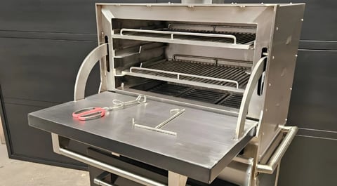 Open stainless steel grill with multiple cooking grates, red handles, and metal shelving on a dark background